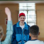 A sports coach in a red cap and blue jacket engages with students in a gym setting.